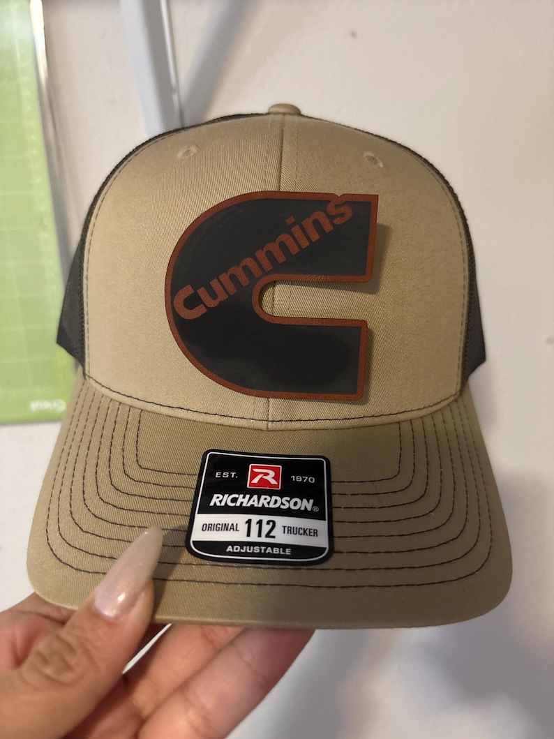 May include: Tan and black trucker hat with the Cummins logo in black and brown. The hat has a curved brim and a mesh back. The Richardson tag is visible on the front. The hat is adjustable.