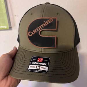 May include: Olive green trucker hat with a black mesh back and a curved brim. The hat features a copper-colored "Cummins" logo on a black background. A black and white tag with the text "RICHARDSON" is attached.