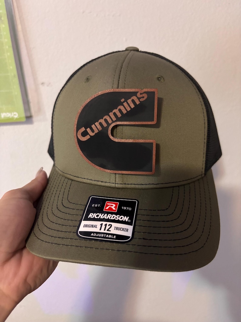 May include: Olive green trucker hat with a black mesh back and a curved brim. The hat features a copper-colored "Cummins" logo on a black background. A black and white tag with the text "RICHARDSON" is attached.