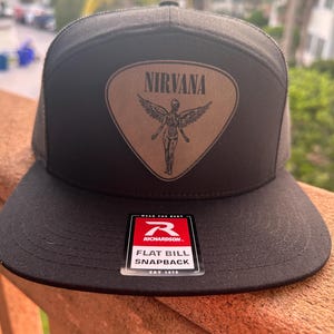 May include: Black flat bill snapback hat with a brown patch featuring the word "NIRVANA" above an angel graphic. The hat has a mesh back and a Richardson tag.