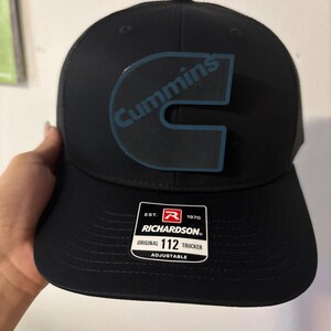 May include: Black trucker hat with the word "Cummins" in blue on the front. The hat has a curved brim and a mesh back. A white tag on the front reads "Richardson Original 112 Trucker Adjustable."
