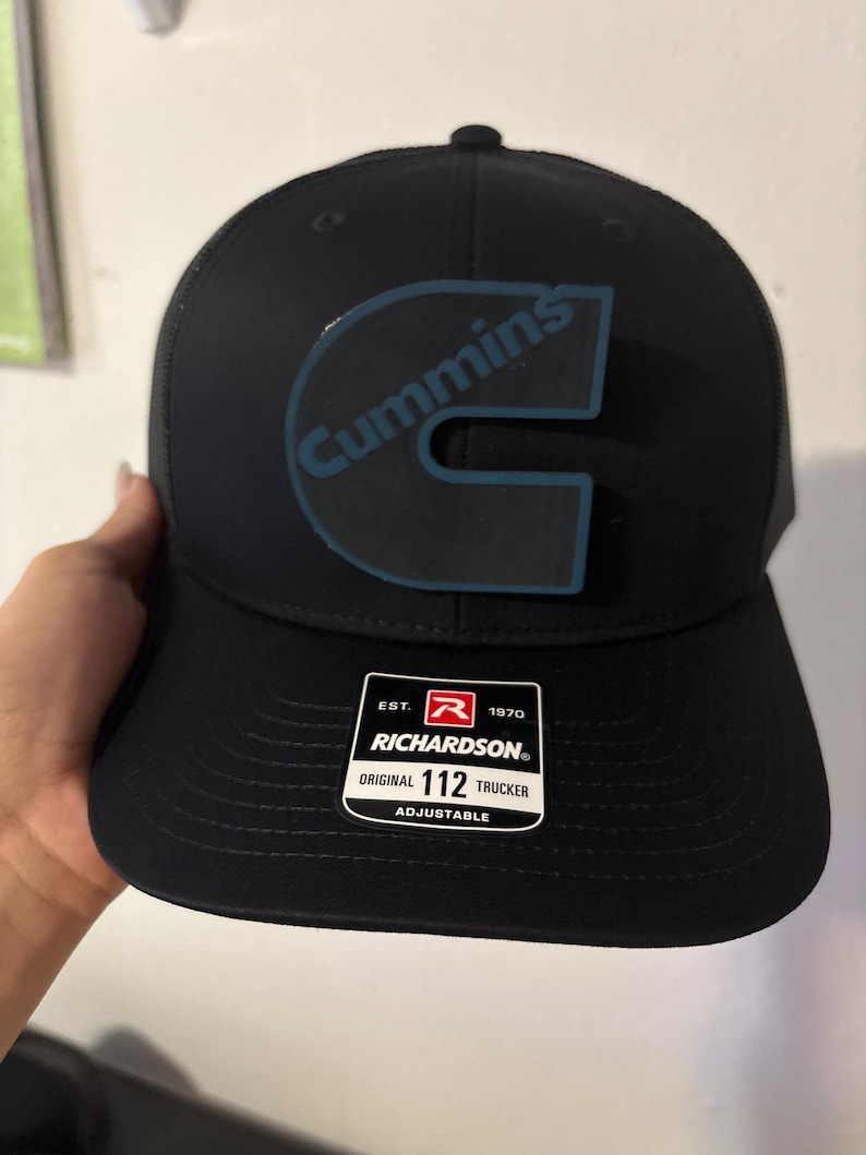 May include: Black trucker hat with the word "Cummins" in blue on the front. The hat has a curved brim and a mesh back. A white tag on the front reads "Richardson Original 112 Trucker Adjustable."