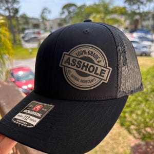 May include: Black trucker hat with a mesh back and a curved brim. The hat features a circular patch with the words "100% Grade A Asshole All Natural Ingredients". The brand tag reads "Richardson 112 Trucker".
