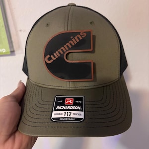 Peut inclure: Casquette de camionneur vert olive et noire avec le logo Cummins en patch noir et marron. La casquette a une visière incurvée et un dos en filet. L'étiquette de la marque Richardson est visible.