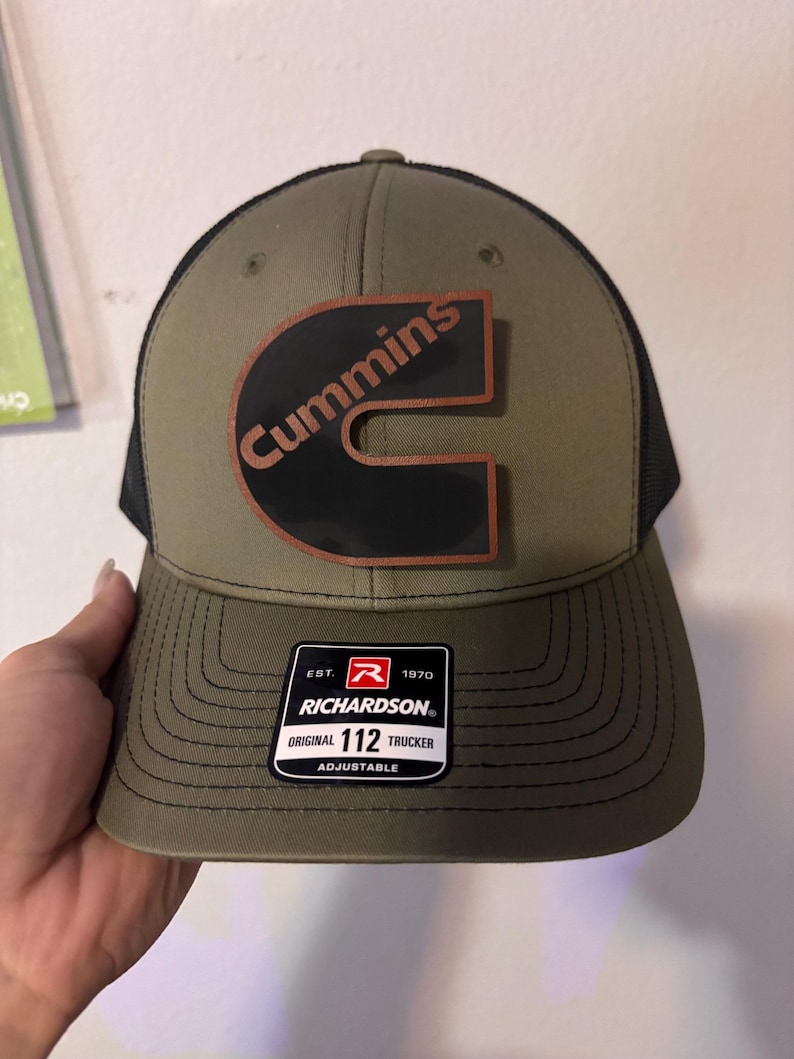 May include: Olive green and black trucker hat with the Cummins logo in a black and brown patch. The hat has a curved brim and a mesh back. The Richardson brand tag is visible.