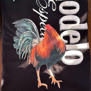 May include: Black t-shirt with a vibrant rooster illustration in shades of red, orange, and teal. The shirt has the word "Modelo" in white and "Especial" in a script font, plus two white bird silhouettes. A casual wear item.