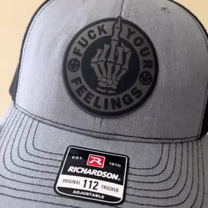 Puede incluir: Gorra de camionero gris con la parte trasera de malla negra y un parche redondo con una mano de esqueleto haciendo la peineta, con las palabras "FUCK YOUR FEELINGS" alrededor. La gorra tiene una visera curva y una etiqueta Richardson 112 Trucker.