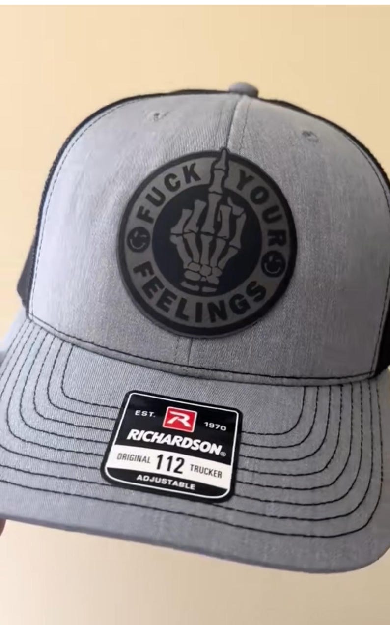 Puede incluir: Gorra de camionero gris con la parte trasera de malla negra y un parche redondo con una mano de esqueleto haciendo la peineta, con las palabras "FUCK YOUR FEELINGS" alrededor. La gorra tiene una visera curva y una etiqueta Richardson 112 Trucker.