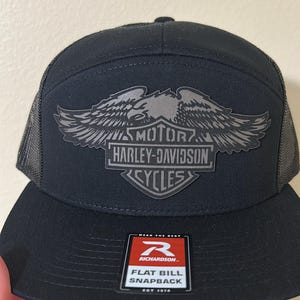 Op de afbeelding: Zwarte flat bill snapback pet met een mesh achterkant. De voorkant heeft een Harley-Davidson logo met een adelaar en de woorden "MOTOR HARLEY-DAVIDSON CYCLES". De pet heeft een Richardson label.