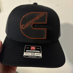 May include: Black Richardson trucker hat with a curved brim and mesh back. The hat features the Cummins logo in orange and the word "Cummins" in orange. The hat has a white tag that says "Richardson Original 112 Trucker Adjustable".