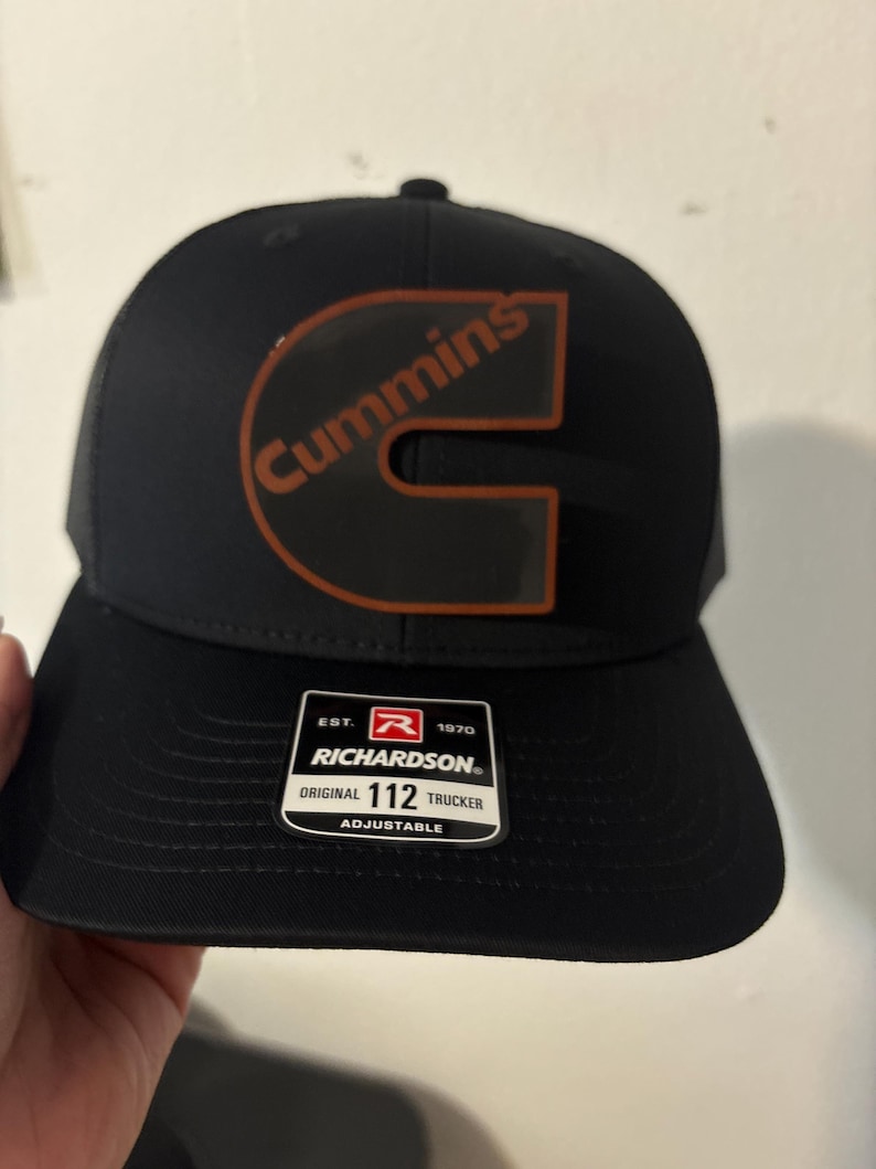 May include: Black Richardson trucker hat with a curved brim and mesh back. The hat features the Cummins logo in orange and the word "Cummins" in orange. The hat has a white tag that says "Richardson Original 112 Trucker Adjustable".