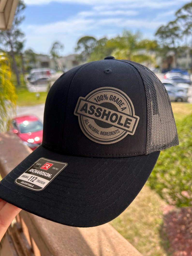 May include: Black trucker hat with a mesh back and a curved brim. The hat features a circular patch with the words "100% Grade A Asshole All Natural Ingredients". The brand tag "Richardson 112 Trucker" is visible.