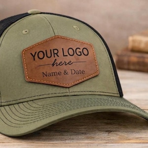 May include: Olive green and black trucker hat with a brown leather patch. The patch features the text "YOUR LOGO here Name & Date". The hat has a curved brim and a mesh back.
