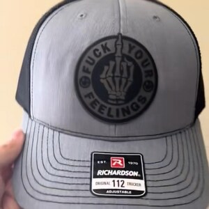 Puede incluir: Gorra trucker gris con parte trasera de malla negra y visera curva. La parte delantera presenta un parche circular con una mano de esqueleto mostrando el dedo medio y las palabras "FUCK YOUR FEELINGS". Con etiqueta Richardson 112 Trucker.