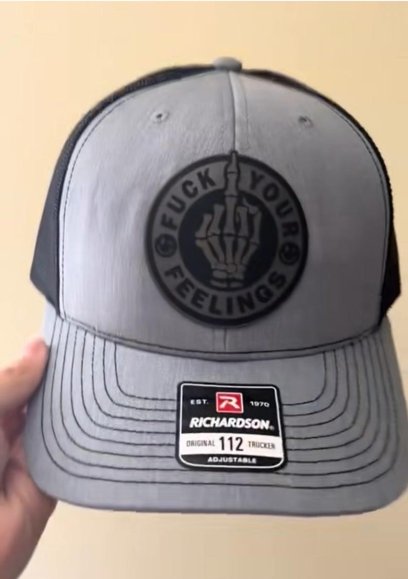 Puede incluir: Gorra trucker gris con parte trasera de malla negra y visera curva. La parte delantera presenta un parche circular con una mano de esqueleto mostrando el dedo medio y las palabras "FUCK YOUR FEELINGS". Con etiqueta Richardson 112 Trucker.