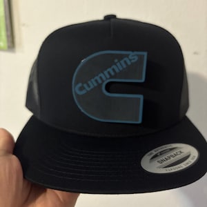 May include: Black snapback hat with the word "Cummins" in light blue lettering. The hat has a black brim and a mesh back. The hat has a "Snapback" sticker on the brim.