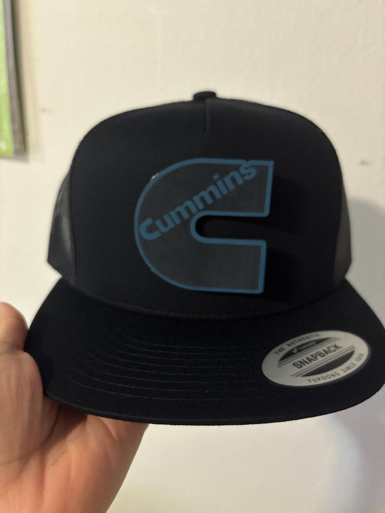 May include: Black snapback hat with the word "Cummins" in light blue lettering. The hat has a black brim and a mesh back. The hat has a "Snapback" sticker on the brim.