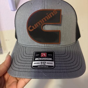 May include: Gray trucker hat with a black mesh back and a curved brim. The hat features the Cummins logo in brown and black, and a Richardson patch. The hat is adjustable.