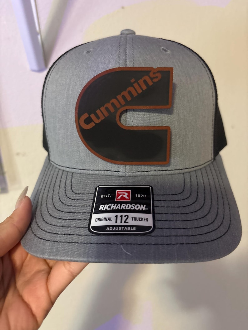 May include: Gray trucker hat with a black mesh back and a curved brim. The hat features the Cummins logo in brown and black, and a Richardson patch. The hat is adjustable.