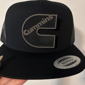 May include: Black trucker hat with a curved brim and mesh back. The hat features a large, gray logo that says "Cummins" in a stylized font. A small, circular sticker is on the brim.