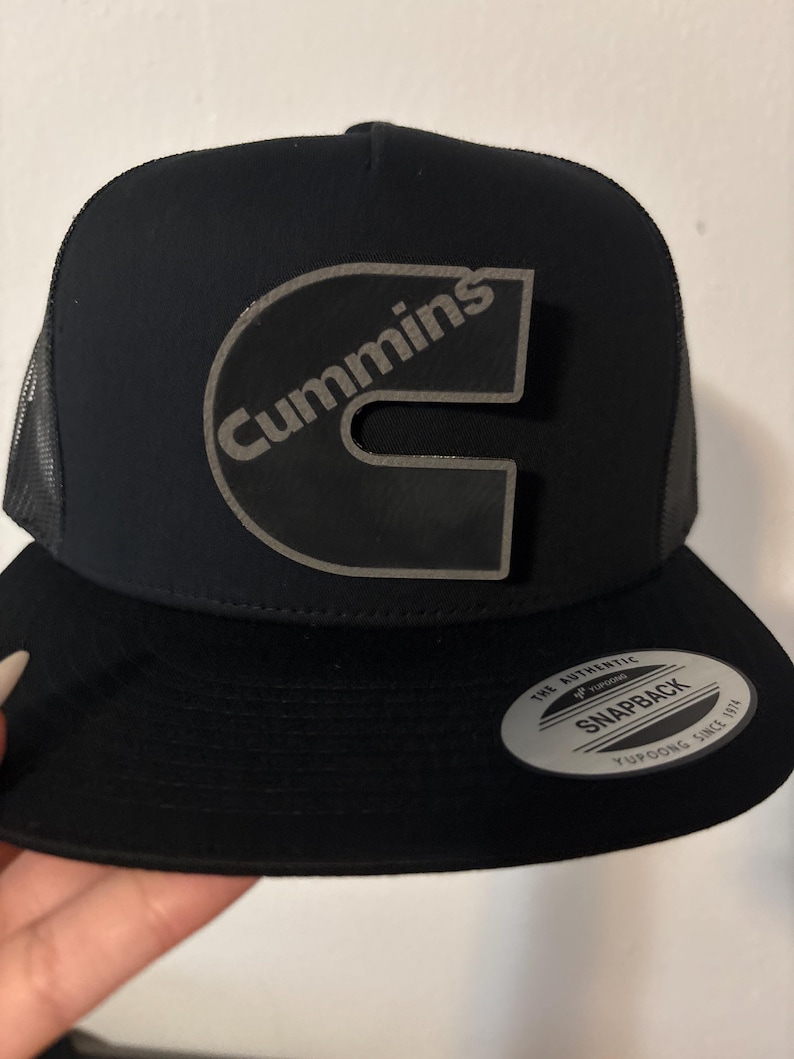 May include: Black trucker hat with a curved brim and mesh back. The hat features a large, gray logo that says "Cummins" in a stylized font. A small, circular sticker is on the brim.