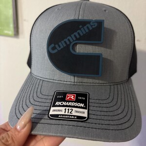 May include: A gray Richardson 112 trucker hat with a black mesh back. The hat features a large black and blue Cummins logo on the front. The hat has a curved brim and an adjustable strap.