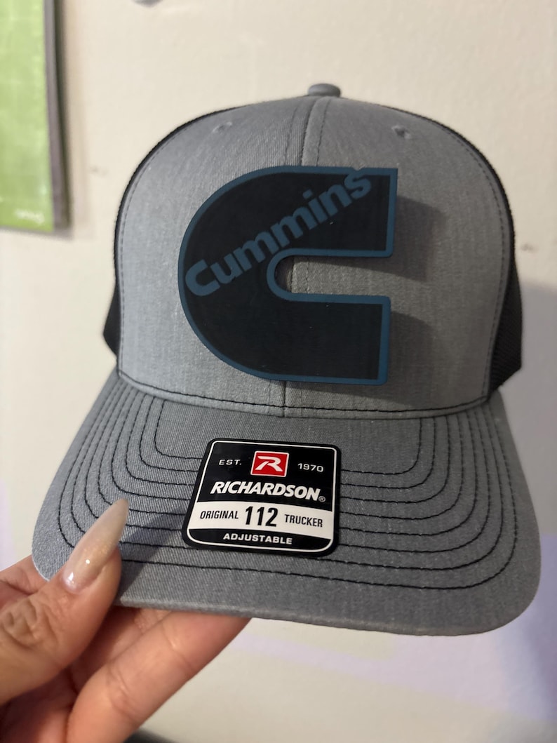 May include: A gray Richardson 112 trucker hat with a black mesh back. The hat features a large black and blue Cummins logo on the front. The hat has a curved brim and an adjustable strap.