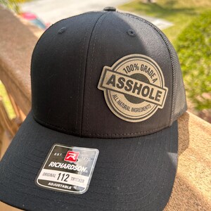 May include: Black trucker hat with a mesh back and a patch that reads "100% Grade A Asshole, All Natural Ingredients." The hat has a curved brim and an adjustable strap. The Richardson brand tag is visible.