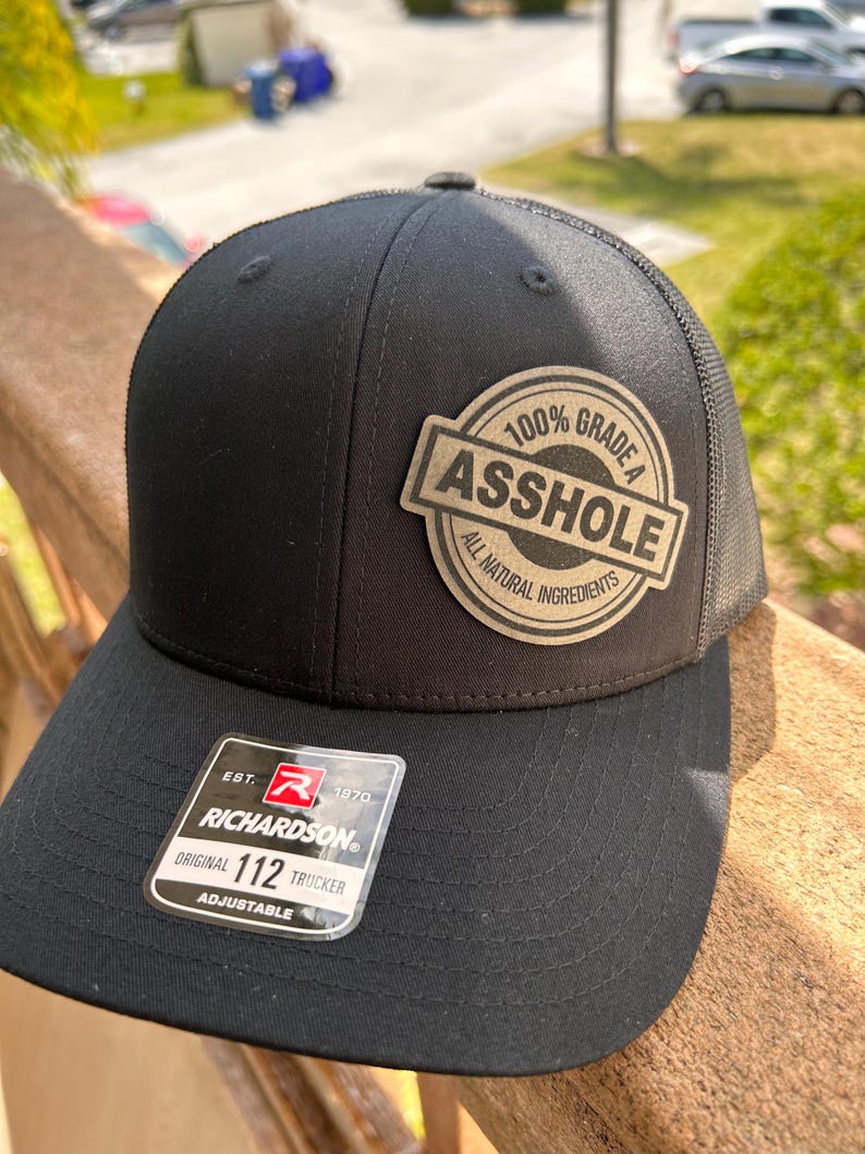 May include: Black trucker hat with a mesh back and a patch that reads "100% Grade A Asshole, All Natural Ingredients." The hat has a curved brim and an adjustable strap. The Richardson brand tag is visible.