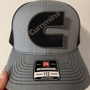 May include: Gray and black trucker hat with the Cummins logo in black. The hat has a curved brim and a mesh back. The Richardson tag reads "Original 112 Trucker Adjustable".