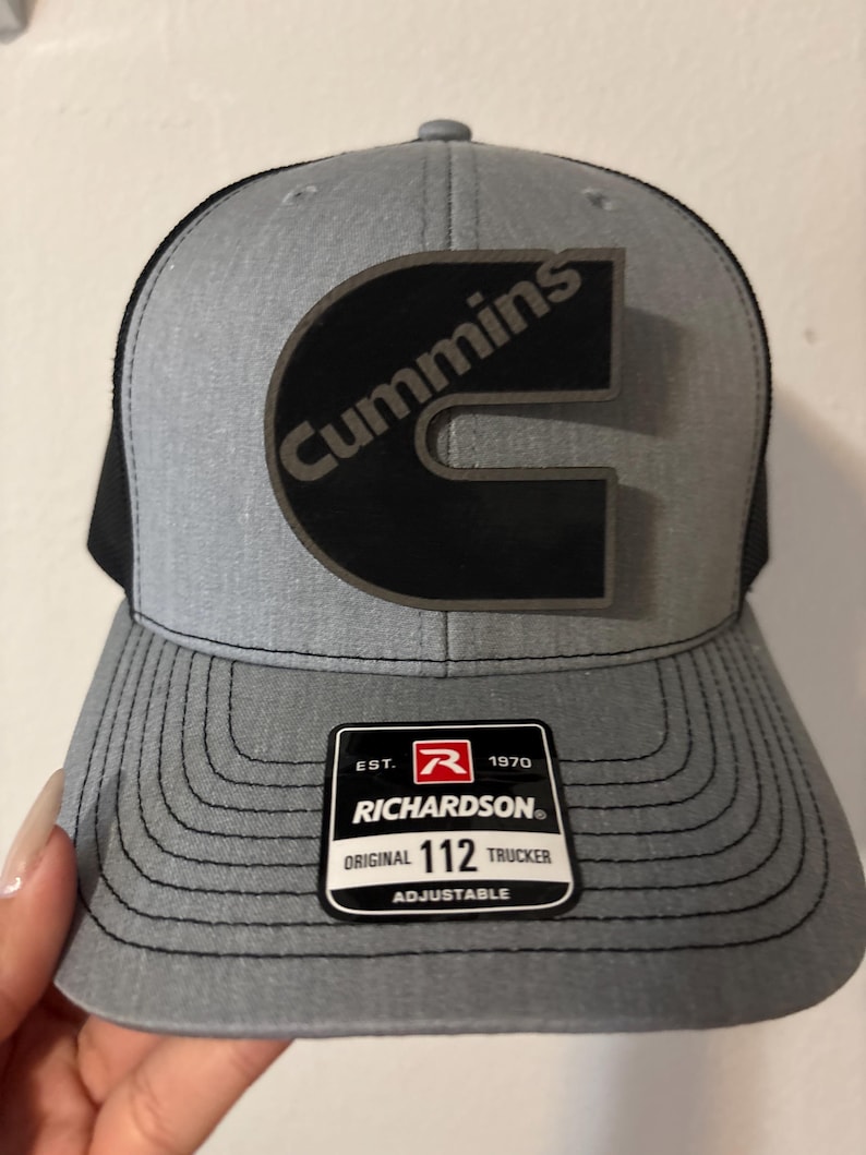 May include: Gray and black trucker hat with the Cummins logo in black. The hat has a curved brim and a mesh back. The Richardson tag reads "Original 112 Trucker Adjustable".