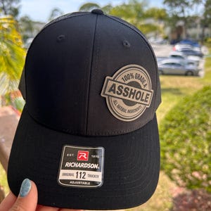 May include: Black trucker hat with a mesh back and a curved brim. The hat features a patch that reads "100% Grade A Asshole, All Natural Ingredients." The Richardson hat is adjustable.