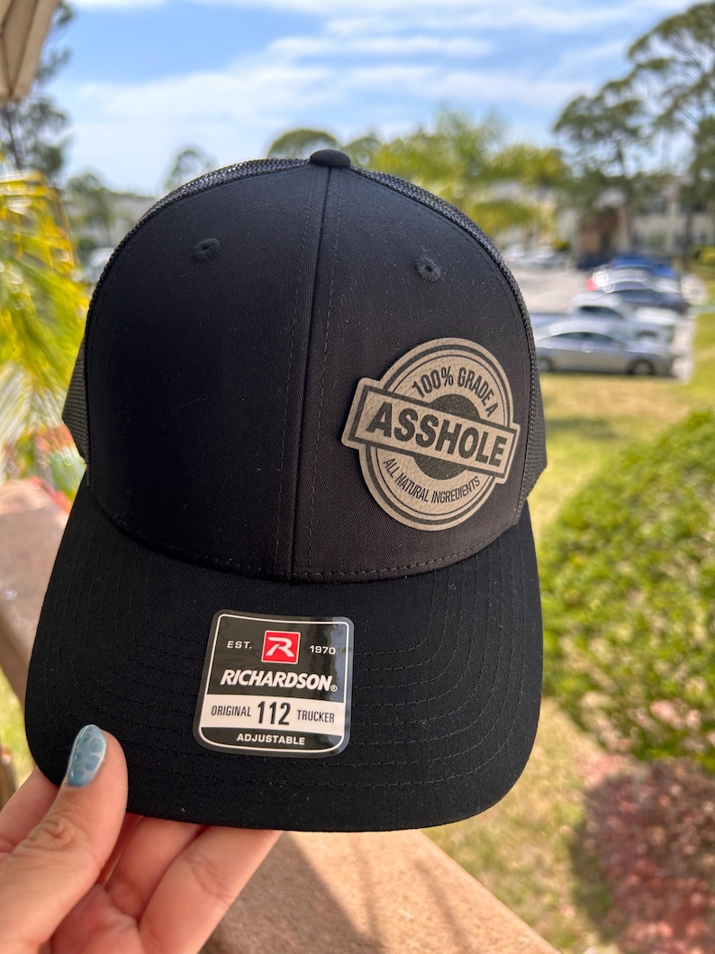 May include: Black trucker hat with a mesh back and a curved brim. The hat features a patch that reads "100% Grade A Asshole, All Natural Ingredients." The Richardson hat is adjustable.