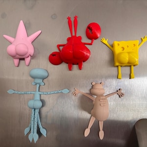 May include: A collection of colorful cartoon character magnets. The magnets include a pink starfish, a red crab, a yellow sponge, a blue squid, and a tan character. Each magnet is a different shape and has a unique design.