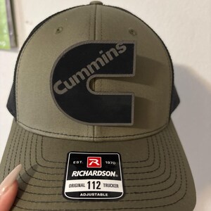 May include: Olive green and black trucker hat with the Cummins logo in black. The hat has a curved brim and a mesh back. The Richardson brand label is visible on the front.