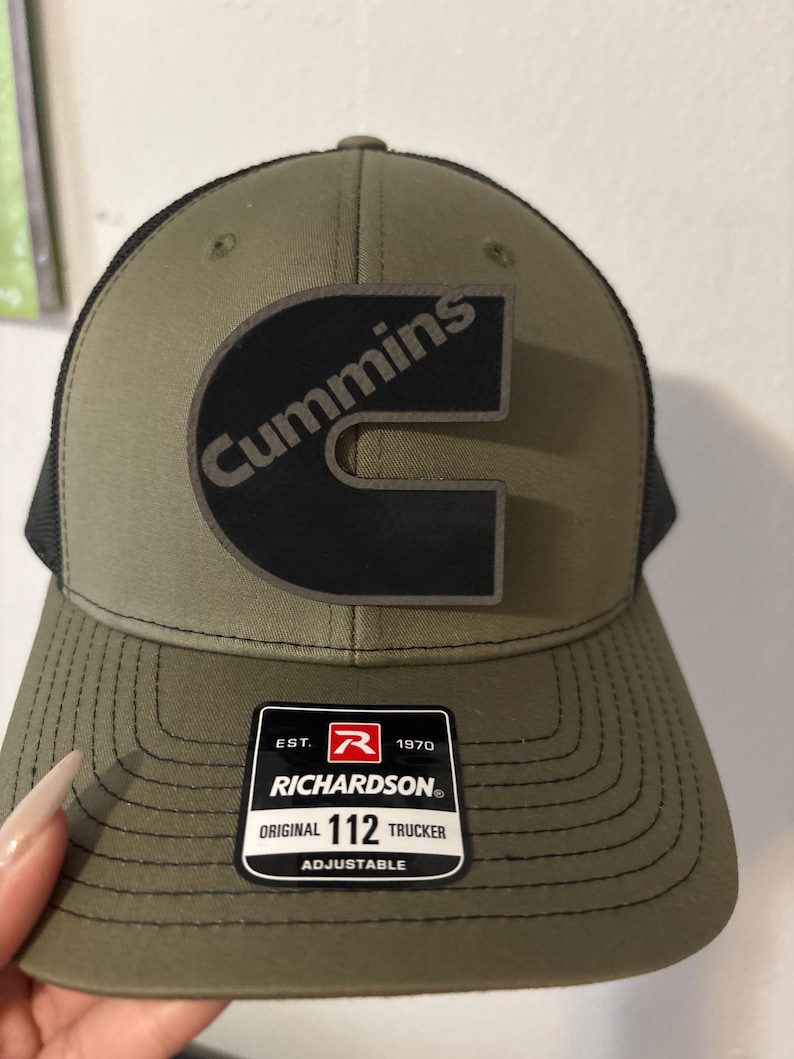 May include: Olive green and black trucker hat with the Cummins logo in black. The hat has a curved brim and a mesh back. The Richardson brand label is visible on the front.