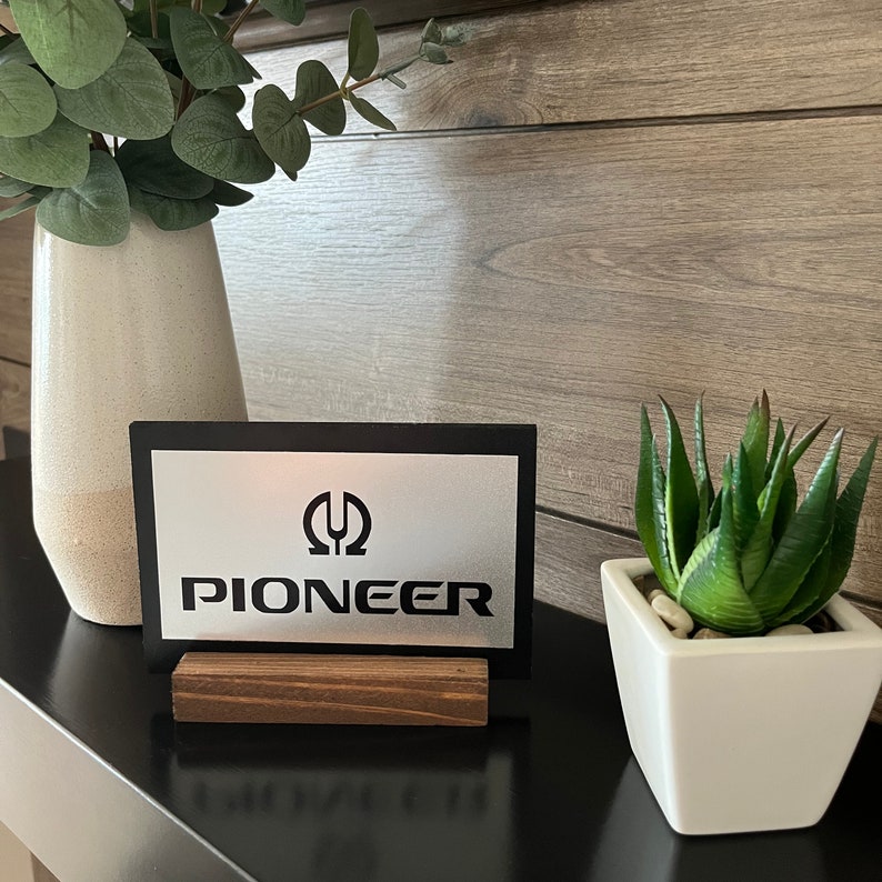 K&ouml;nnte beinhalten: Ein schwarzes Schild mit silbernem Hintergrund und dem Text "Pioneer" sowie einem stilisierten Logo. Das Schild steht auf einem Holzst&auml;nder und befindet sich auf einer Holzoberfl&auml;che.