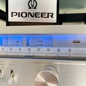 K&ouml;nnte beinhalten: Ein silberner Pioneer Stereo-Receiver mit einem silbernen Knopf und einem schwarzen und silbernen Pioneer-Logo. Der Receiver hat ein blau beleuchtetes Display mit den Worten "SIGNAL" und "TUNING" und Zahlen von 94 bis 108.