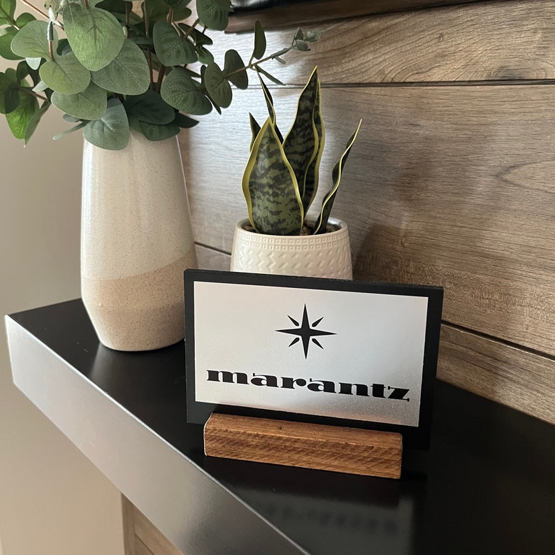 Puede incluir: Un letrero negro y plateado con la palabra "marantz" y un logotipo de estrella en un soporte de madera. El letrero est&aacute; sobre un estante negro.