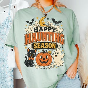 Pode incluir: Uma t-shirt verde-clara com uma estampa gráfica contendo o texto "HAPPY HAUNTING SEASON" em laranja e preto. O design inclui uma lua crescente amarela, dois morcegos pretos, um gato preto com uma gravata borboleta laranja, uma abóbora e um fantasma.
