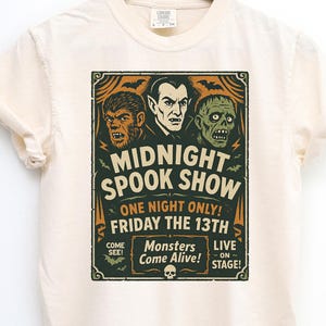 May include: Ivory t-shirt with a vintage "Midnight Spook Show" design. The graphic features a werewolf, Dracula, and a zombie, with the text "One Night Only! Friday the 13th." The shirt is hanging on a wooden hanger.
