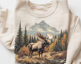 Peut inclure: Un sweat-shirt couleur sable avec une illustration détaillée d'un orignal dans une forêt avec une montagne en arrière-plan. Le motif comprend des arbres, un feuillage d'automne et un petit ruisseau. Les mots "Color Sand" sont écrits en cursive.