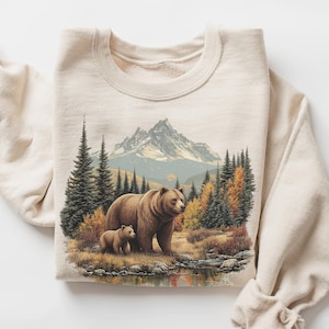 May include: A sand-colored sweatshirt featuring a detailed illustration of a bear and cub in a forest scene with a mountain backdrop. The design includes trees, a lake, and autumn foliage. The text "Color Sand" is in the bottom right corner.