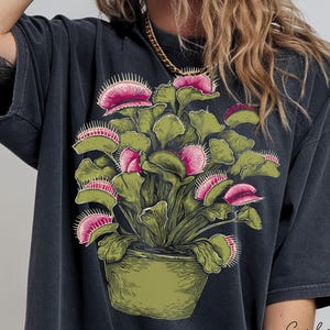 May include: Black t-shirt with a detailed illustration of a Venus flytrap plant in a green pot. The plant has green leaves and pink and white traps. The words "Color" and "Black" are printed on the bottom right.