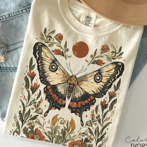 May include: Ivory-colored t-shirt featuring a large butterfly illustration with intricate details in black, brown, and orange. The design includes floral elements and a sun graphic. The text "Color IVORY" is printed on the bottom right.