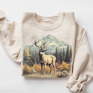 May include: A sand-colored sweatshirt featuring a detailed graphic of a deer in a forest with a mountain backdrop. The deer has large antlers and is standing in a field of grass and bushes. The sweatshirt is folded with the sleeves rolled up.