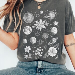 Puede incluir: Una camiseta gris oscuro con un estampado blanco de planetas, estrellas y flores. El texto "Non Obeam" es visible en la manga derecha.