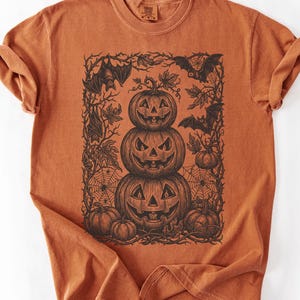 Könnte beinhalten: Ein kurzärmeliges T-Shirt in gebranntem Orange mit einem schwarzen Grafikdruck im Tintenstil. Das Motiv zeigt einen Stapel aus drei geschnitzten Kürbissen, umgeben von einem Rahmen aus verschlungenen Ranken, Herbstblättern und Fledermäusen.