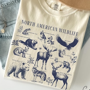 Puede incluir: Una camiseta de color crema con un gráfico azul de vida silvestre de América del Norte. El gráfico incluye animales como un oso, un alce, un lobo y un águila. El texto "North American Wildlife" está impreso en azul en la parte superior del gráfico. La camiseta está colocada sobre un fondo de mezclilla azul.
