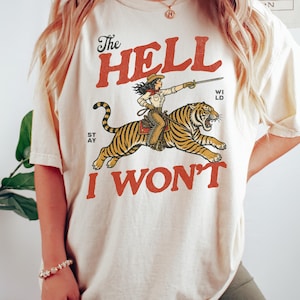 以下が含まれることがあります： 女性がトラに乗っているグラフィックが特徴のアイボリー色のTシャツ。赤で「The Hell I Won't」と書かれています。このシャツは半袖でゆったりとしたフィット感です。色は「Ivory」です。